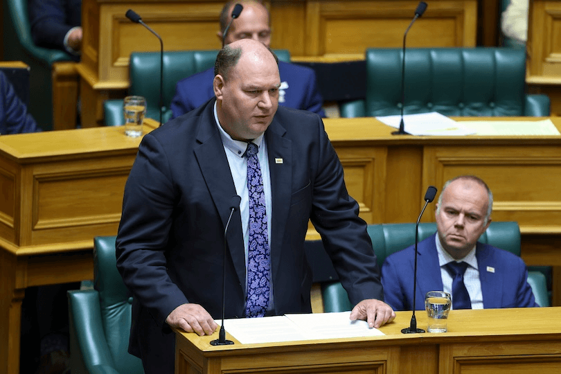 A man in a suit stands at a podium speaking in a parliamentary chamber, with another man seated nearby and several others partly visible in the background.