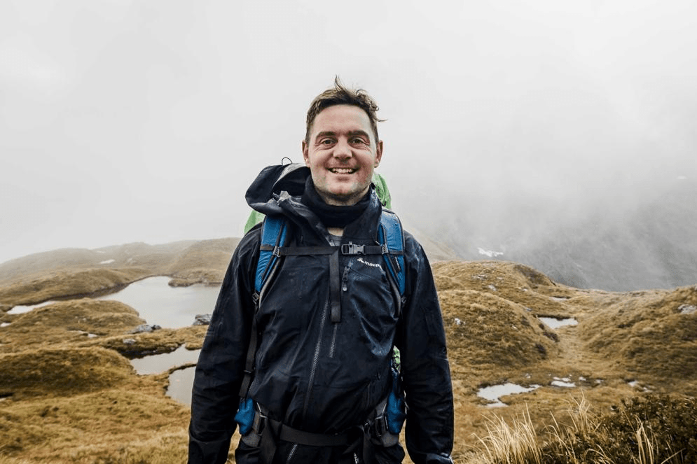 A person wearing a black rain jacket and backpack smiles while standing outdoors in a foggy, grassy landscape with small ponds and rolling hills in the background.