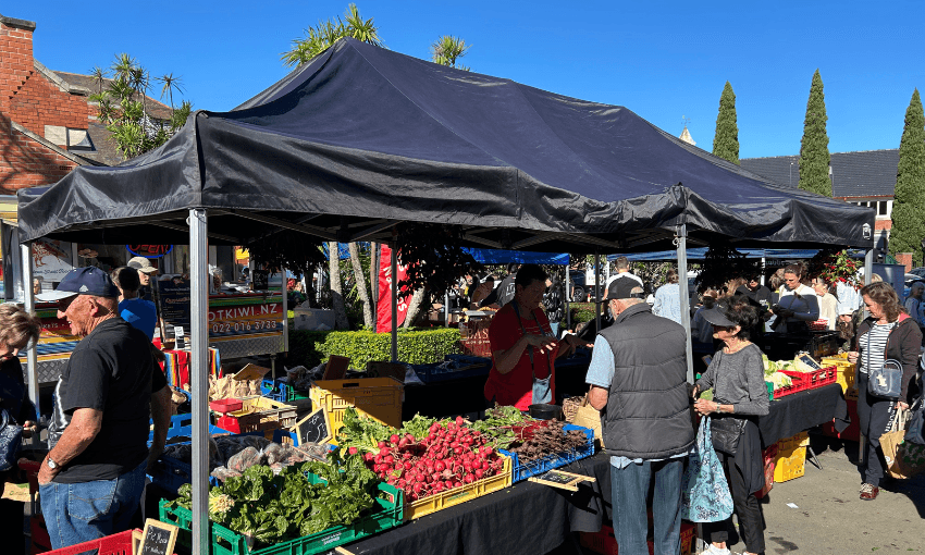 People shop for fresh produce at an outdoor market stall under a large black canopy on a sunny day. Various vegetables and fruits are displayed in crates on the tables, and trees and buildings are visible in the background.