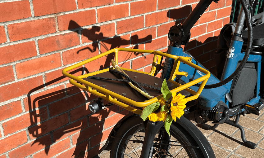 A yellow-front basket on a blue bicycle holds sunglasses and is decorated with a bright sunflower, parked against a sunlit red brick wall.