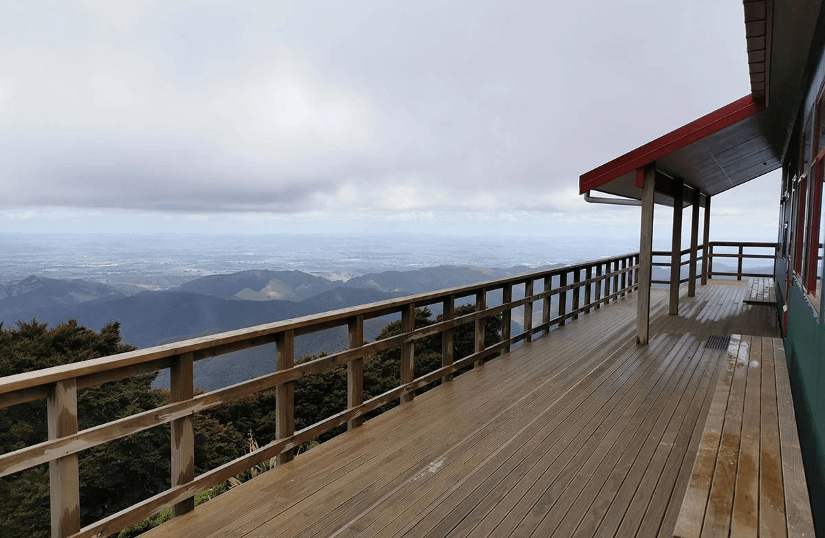 A landscape view from a large wooden deck showing a long railing and grassy hills below a cloudy sky