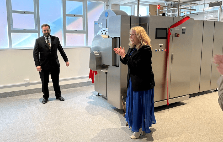 A man in a suit and a woman in a blue dress clap hands next to a large silver machine