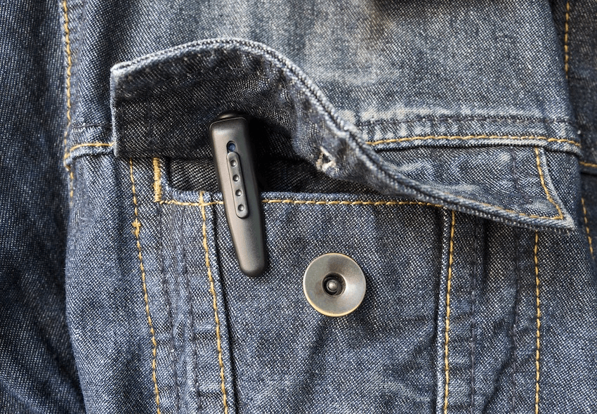 A small black spy camera hidden in a pen clipped to the pocket of a blue denim jacket with a visible button and detailed stitching.