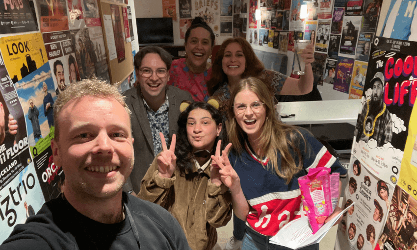 A group of six people smile and pose for a selfie in a hallway covered with colorful event posters. One person is holding up a peace sign, and another is holding a snack bag and a drink. The atmosphere is cheerful and lively.