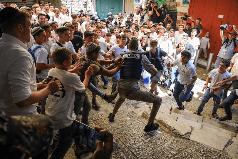 A large group of young men confront a uniformed journalist wearing a "Press" vest on a stone street. Some appear agitated, with raised arms, while others look on. Photographers and onlookers observe the tense scene.