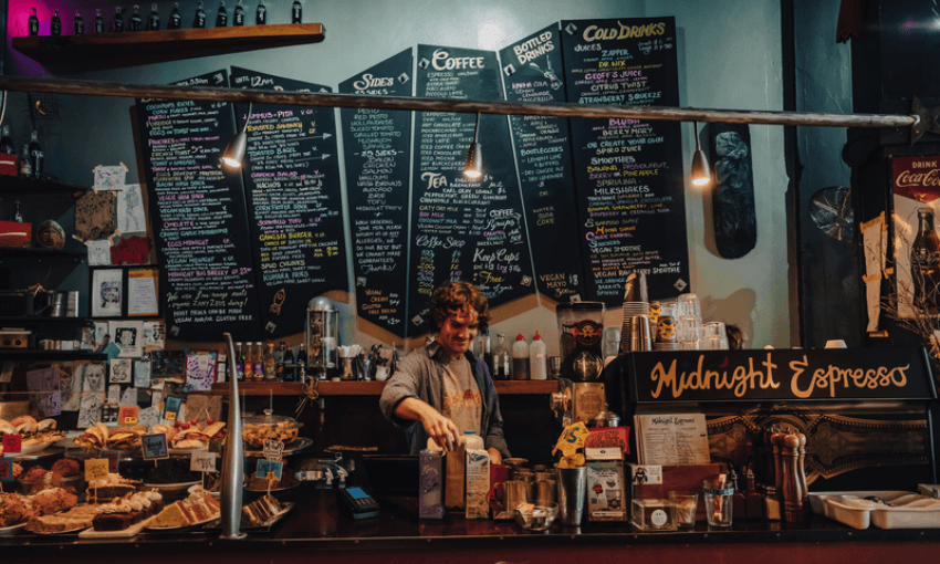 A barista stands behind the counter at a cozy café called "Midnight Espresso," with pastries on display and a large menu with colorful chalk writing hanging on the wall behind them.
