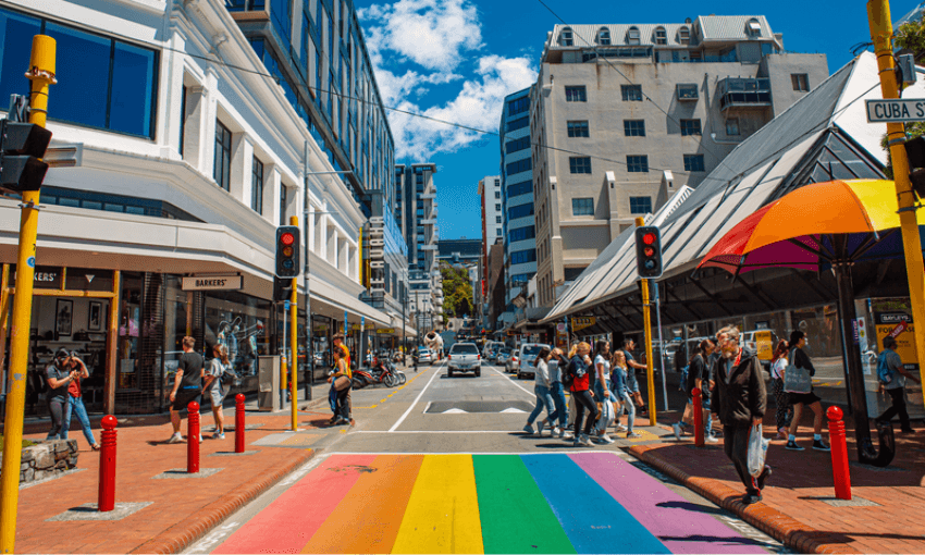 A vibrant rainbow-colored crosswalk spans a busy city street lined with modern buildings. Pedestrians wait and cross under bright sunshine, with shops and a yellow-and-orange umbrella visible. Traffic lights are red.