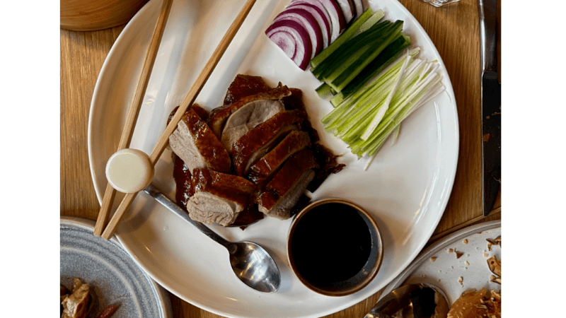 A plate with sliced roast duck, hoisin sauce, cucumber sticks, sliced scallions, and red onion, with wooden chopsticks and a spoon, set on a wooden table.