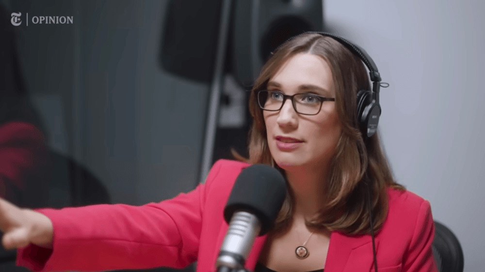 A woman wearing glasses and headphones speaks into a microphone in a recording studio. She is wearing a pink blazer and gesturing with her hand.