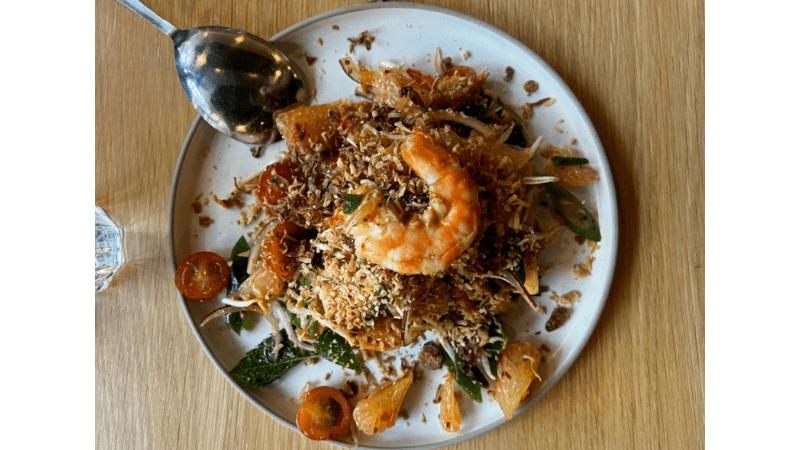 A plate of salad topped with a large shrimp, shredded vegetables, cherry tomatoes, and crispy toppings is served on a wooden table next to a metal spoon.