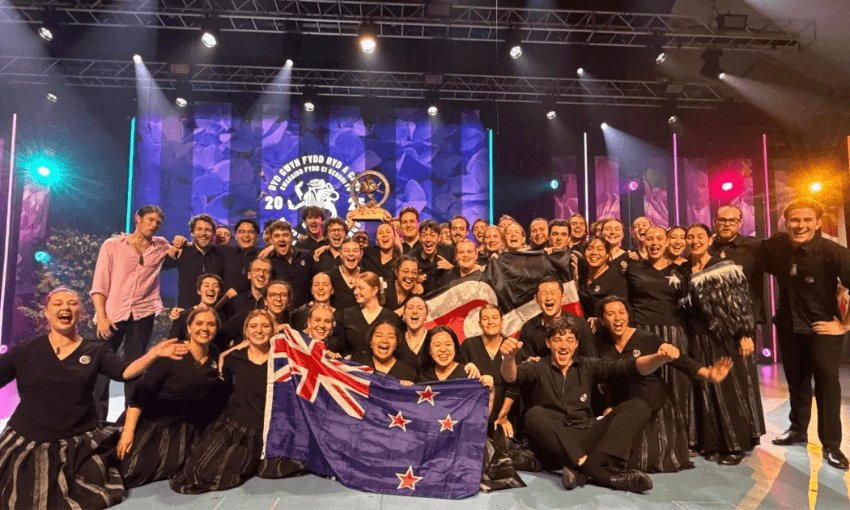 A group photo of the 2025 youth choir. About 40 young people wearing black and silver uniforms and smiling while holding a trophy and a New Zealand flag