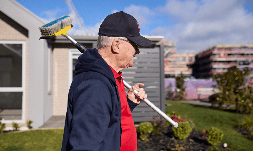 An older man wearing a cap and glasses carries a broom over his shoulder outside a modern house on a sunny day, with construction buildings visible in the background.