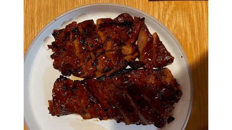 A plate with two portions of sliced, glazed barbecued pork, featuring a shiny, caramelized exterior and slight charring, set on a light-colored plate atop a wooden table.