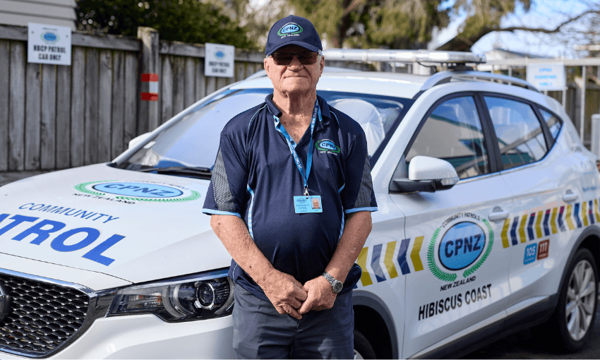 An older man in a navy CPNZ uniform and cap stands in front of a Community Patrol car marked "Hibiscus Coast" in a parking lot, wearing sunglasses and an ID badge.
