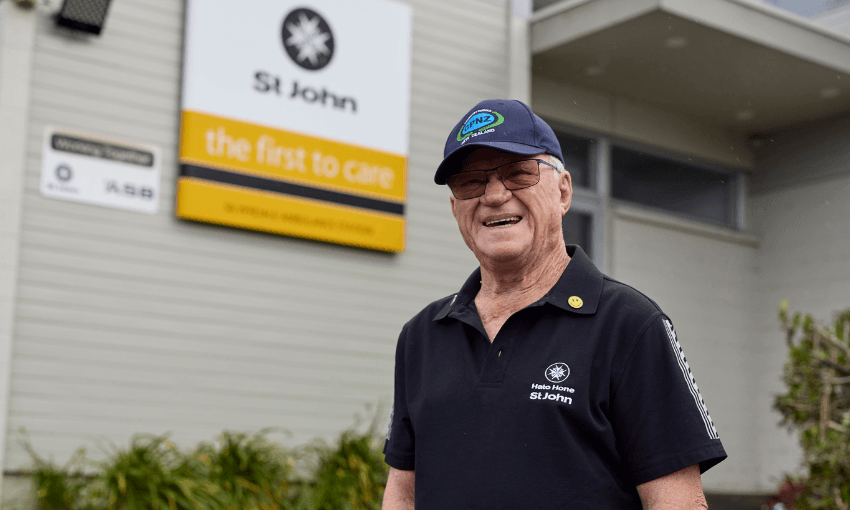 An older man wearing a St John uniform and cap smiles outside a building with a St John sign that reads "the first to care." Green plants are visible near the building’s entrance.