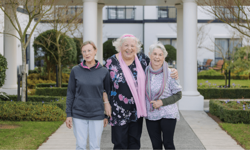 Three older women stand arm in arm and smiling outside a building, surrounded by greenery and trees on a pathway. They are dressed casually and appear happy and relaxed, enjoying each other's company.