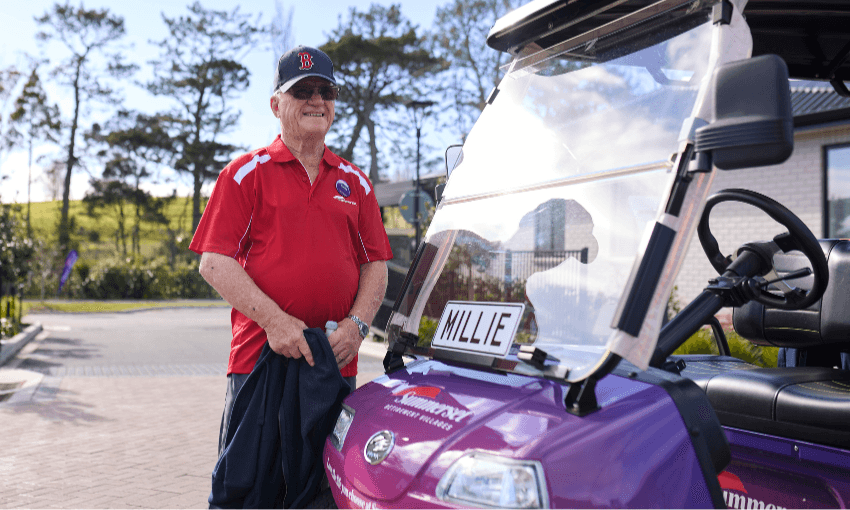 An older man in a red shirt and cap smiles while standing beside a purple golf cart labeled "MILLIE" on a sunny day, with trees and a building in the background.