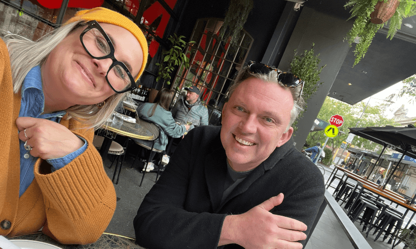 Two smiling adults sit at an outdoor cafe table. The woman wears a mustard beanie, glasses, and a tan sweater; the man wears sunglasses on his head and a dark jacket. Other people and greenery are visible in the background.