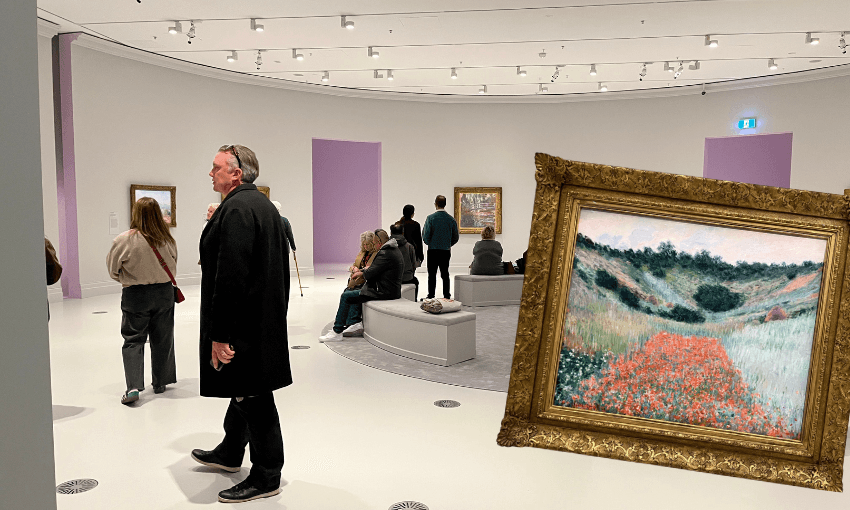 People view paintings in a modern, curved art gallery with white walls and lavender accents; a framed landscape painting of a flower-filled field is highlighted in the foreground.
