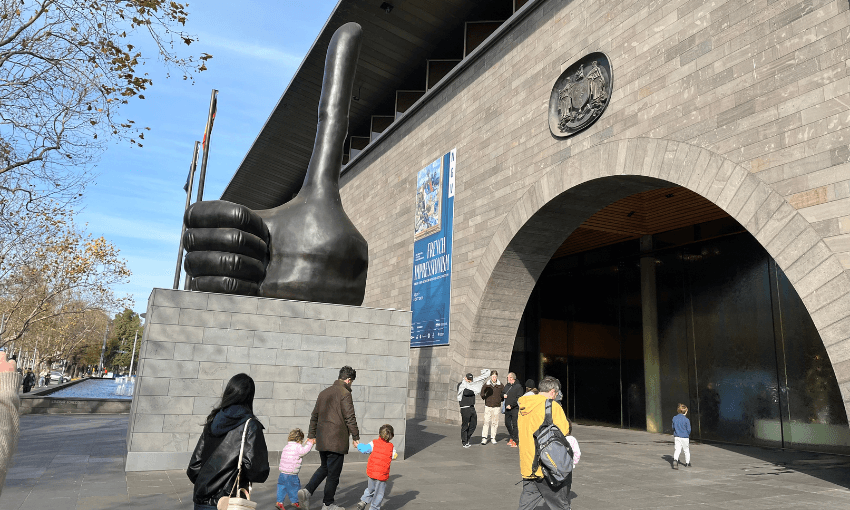 People walk past a large outdoor sculpture of a hand giving a thumbs up near the entrance of a stone building on a sunny day. Some children and adults are gathered around the sculpture.