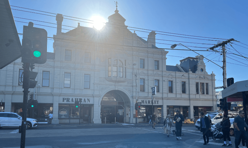 People cross a busy street in front of the historic Prahran Market building in bright sunlight, with cars waiting at the intersection and the sun shining from behind the ornate facade.