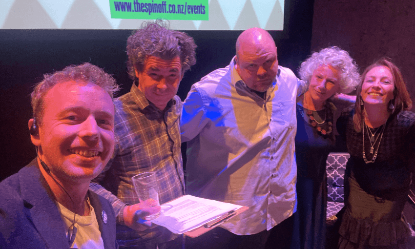 Five people stand closely together, smiling at the camera under purple lighting. One person in front holds a clipboard and a drink. A screen with partially visible text is in the background.