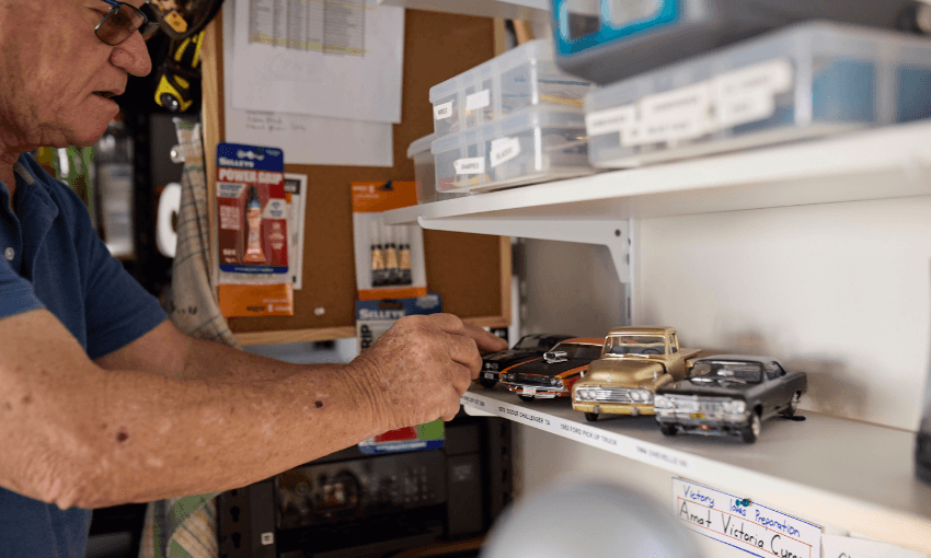 An man arranges vintage model cars on a white shelf in a workshop area, with labeled boxes, tools, and various supplies organized in the background.