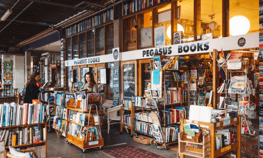 A cozy bookstore called Pegasus Books has bookshelves and carts filled with colorful books outside. Two people browse among the selections, and large windows reveal even more books inside the warm, inviting shop.
