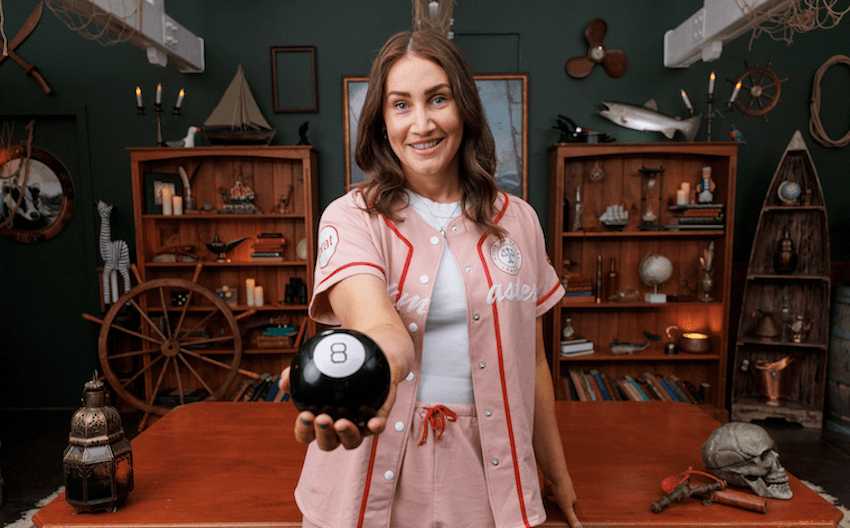 Bree wears a baseball outfit with long brown hair and holds a number eight ball towards the camera. 