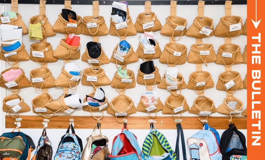 Wicker baskets holding various personal items are hung in rows on a wall, each labeled with a name tag. Below, colorful children's backpacks are hanging on hooks. The words "THE BULLETIN" appear in orange on the right side.