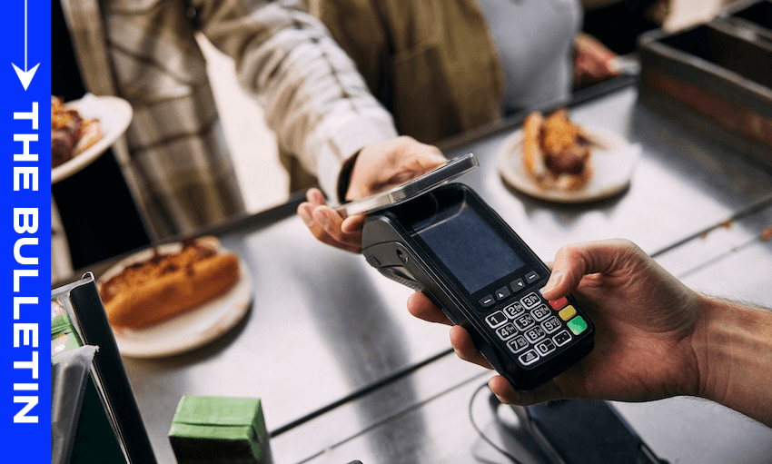 A person holds a smartphone over a payment terminal to make a contactless payment at a food counter, with plates of food visible in the background. A blue banner on the side reads "THE BULLETIN.