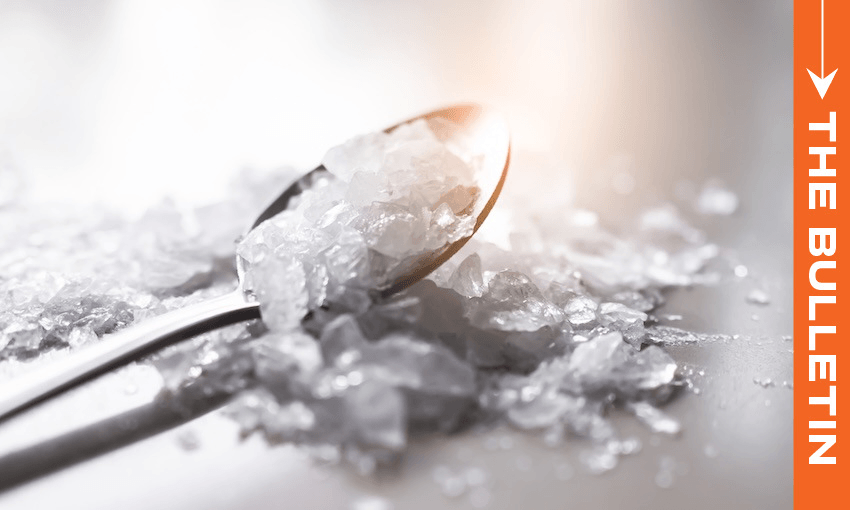 A close-up of a metal spoon holding clear, crystalline chunks, scattered on a shiny surface. An orange vertical banner on the right reads “THE BULLETIN.”.