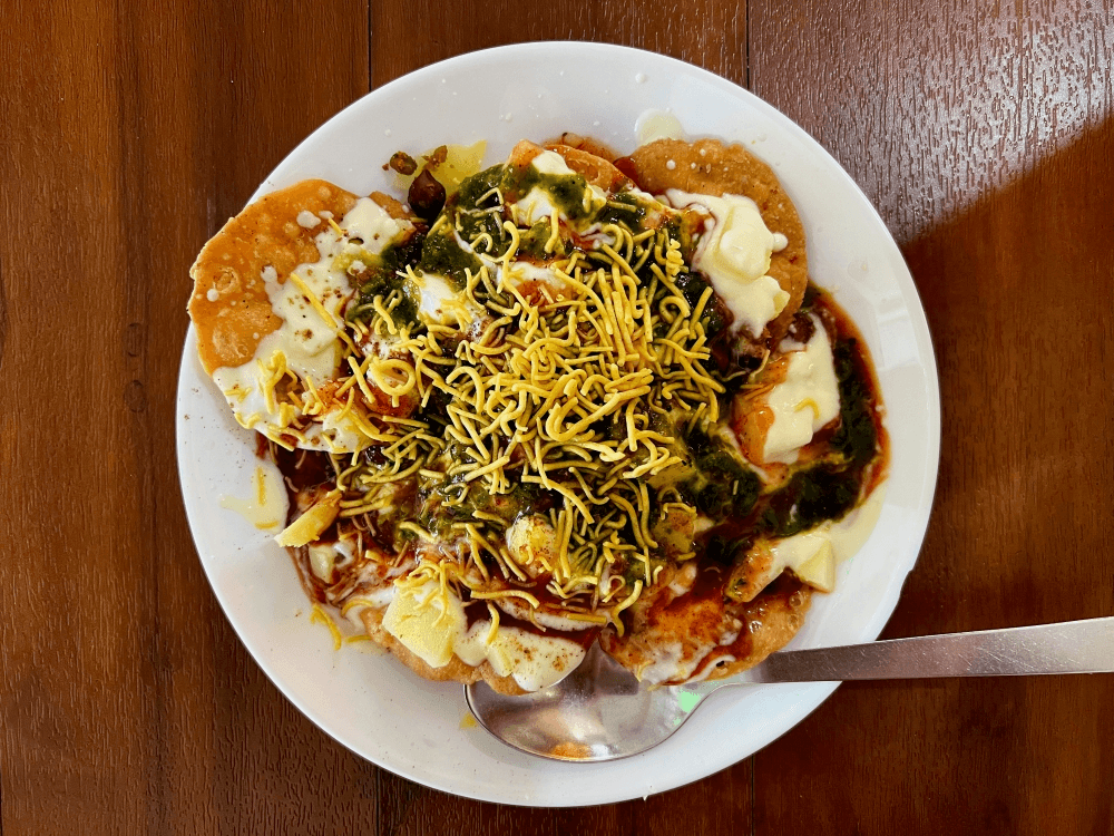A plate of Indian chaat topped with crispy sev, yogurt, chutneys, and diced potatoes, served with a spoon on a wooden table.