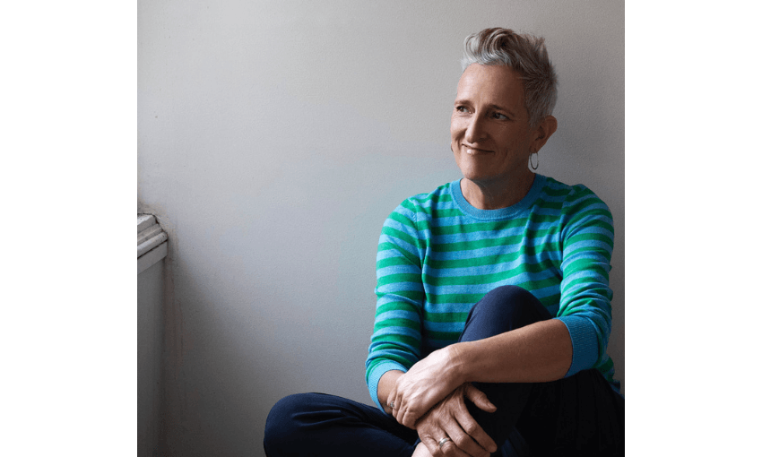 Photo of a woman sitting against a wall, smiling. She has grey hair and is wearing a green striped top.
