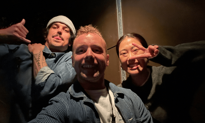 Three people pose for a selfie in dim lighting. The person on the left flashes a shaka sign, the person in the center smiles at the camera, and the person on the right makes a peace sign with their fingers.