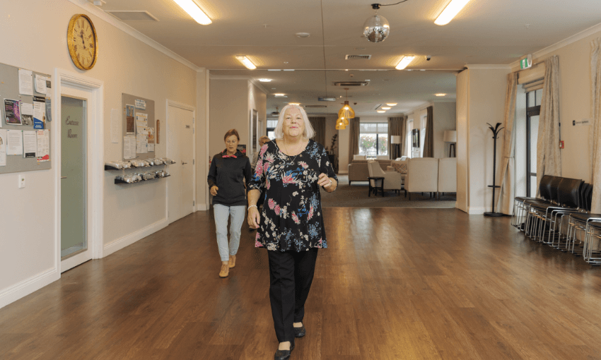 Two women walk down a spacious, well-lit hallway with wooden floors and beige walls. One woman is in the foreground, wearing a floral top, while the other follows behind. The area has chairs, a clock, and a bulletin board.