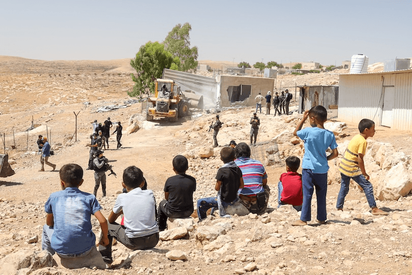 A group of children sit and stand on rocky ground watching as armed soldiers and a bulldozer approach a metal building in a dry, rural area. Other people observe the scene, and several buildings are visible in the background.