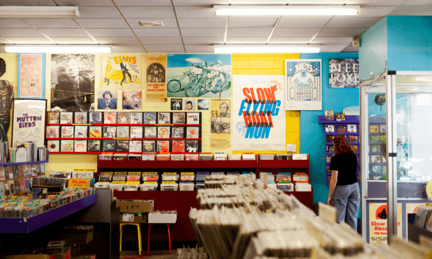 A person stands inside a colorful record store, browsing shelves of vinyl records. The walls are covered with vibrant posters and album covers, creating a retro and eclectic atmosphere.