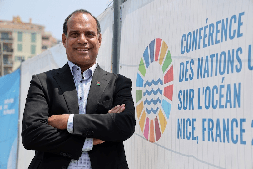 A man in a black suit stands smiling with arms crossed in front of a fence displaying the United Nations Ocean Conference logo and text reading “Conférence des Nations Unies sur l’Océan, Nice, France.”.