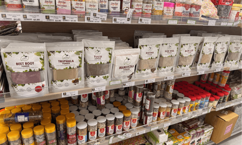 Packets of herbal powders, including beetroot, triphala, tulsi, and others, are displayed on a shelf in a store. Below are jars of various herbs and spices in yellow and red lids.