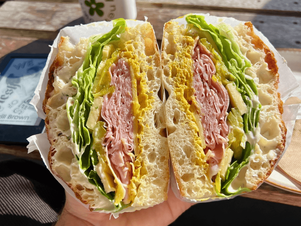 A hand holds a large sandwich cut in half, revealing layers of lettuce, sliced meats, cheese, pickles, and mayonnaise on crusty bread. Sunlight highlights the fresh ingredients.