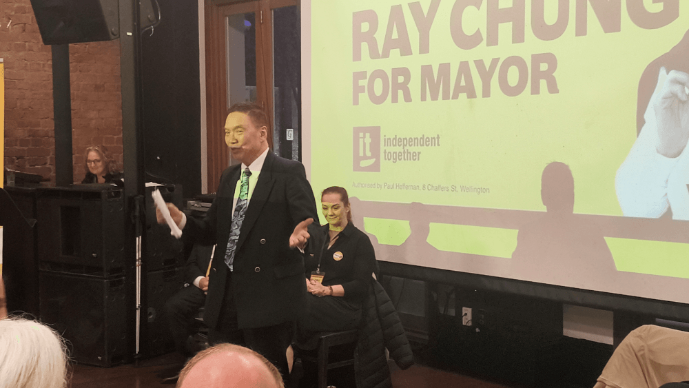 A man in a suit speaks in front of an audience at an indoor event. Behind him, a large yellow screen displays the text “Ray Chung for Mayor” and “independent together.” Several people are seated nearby.