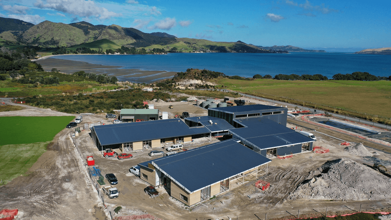 Aerial view of a coastal construction site with multiple buildings, vehicles, green hills, and water in the background under a blue sky with scattered clouds.