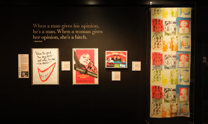 A museum display featuring vintage posters, a colorful pop art panel on the right, and a quote on the wall that reads: "When a man gives his opinion, he's a man. When a woman gives her opinion, she's a bitch.