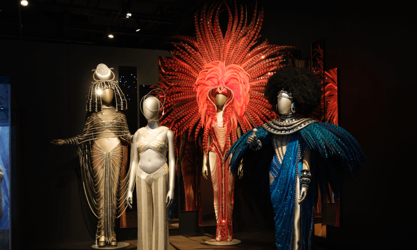 Four mannequins display elaborate show costumes, including gold gowns, a blue feathered outfit with an afro wig, and a striking pink costume with a large feathered headdress, set against a dark exhibit backdrop.