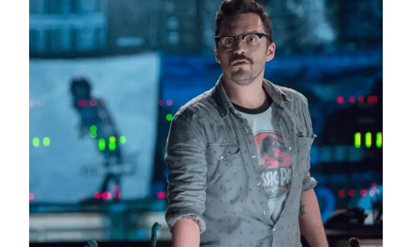 A still from Jurassic World showing the actor Jake Johnson who is a young man with a moustache and glasses. He is in a control room and is wearing a Jurassic Park T-shirt.