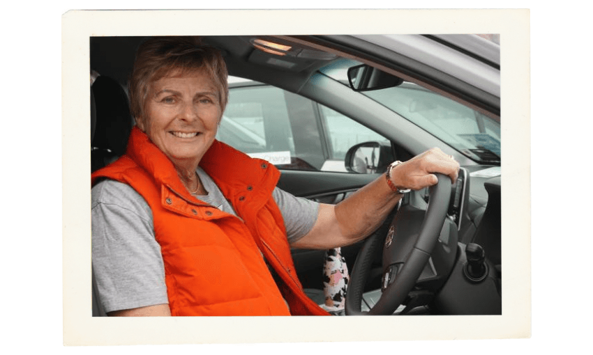 A woman wearing an orange vest and gray shirt smiles while sitting in the driver’s seat of a car, holding the steering wheel with one hand.