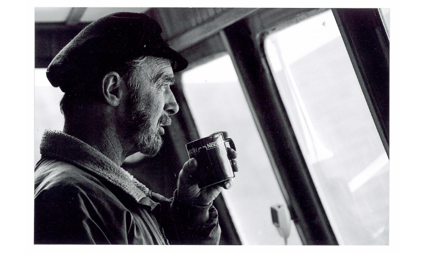 Black and white photo of a man staring out a window about to sip a mug of hot drink.