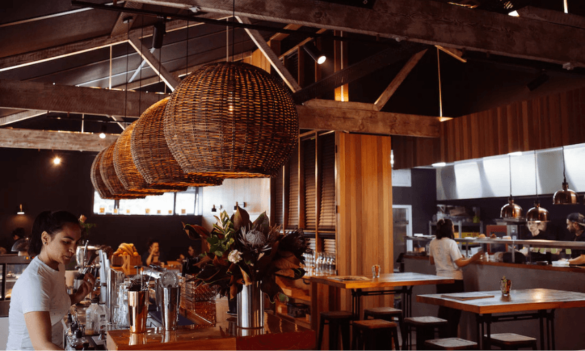 A stylish, modern restaurant interior with wooden beams, wicker pendant lights, and an open kitchen. A bartender prepares drinks at the counter, while other staff and diners sit at tables in the warm, inviting space.