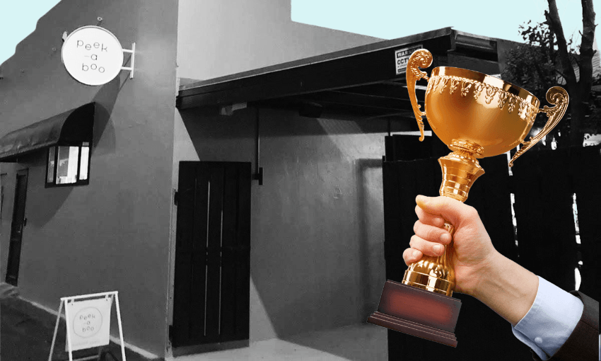 A hand holding a gold trophy in front of a building with a round "Peek-a-boo" sign; the image is mostly black and white except for the trophy, which is in color.